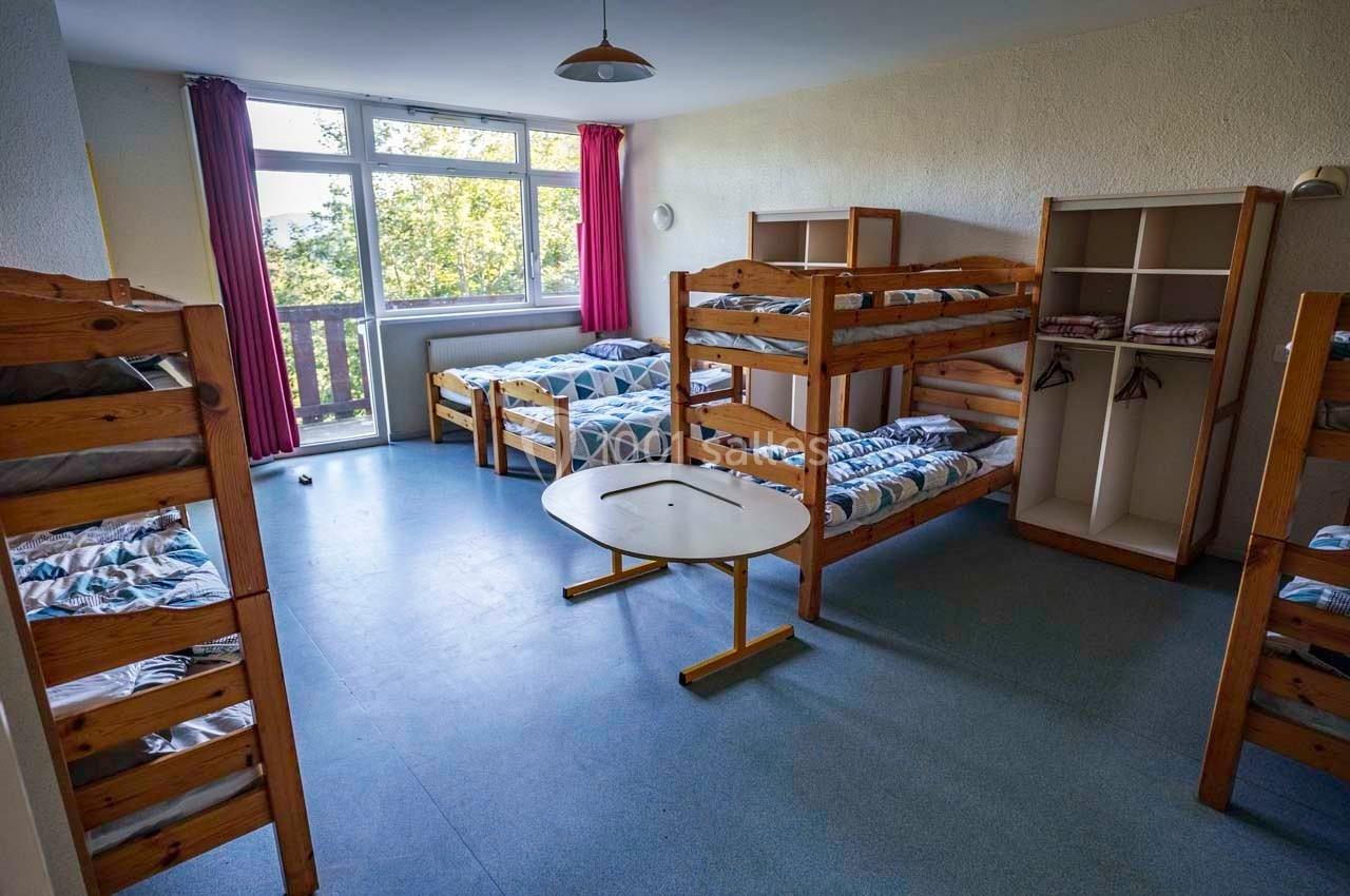 Dortoir avec lits superposés en bois, fenêtres donnant sur l'extérieur, armoires ouvertes et sol en lino bleu.