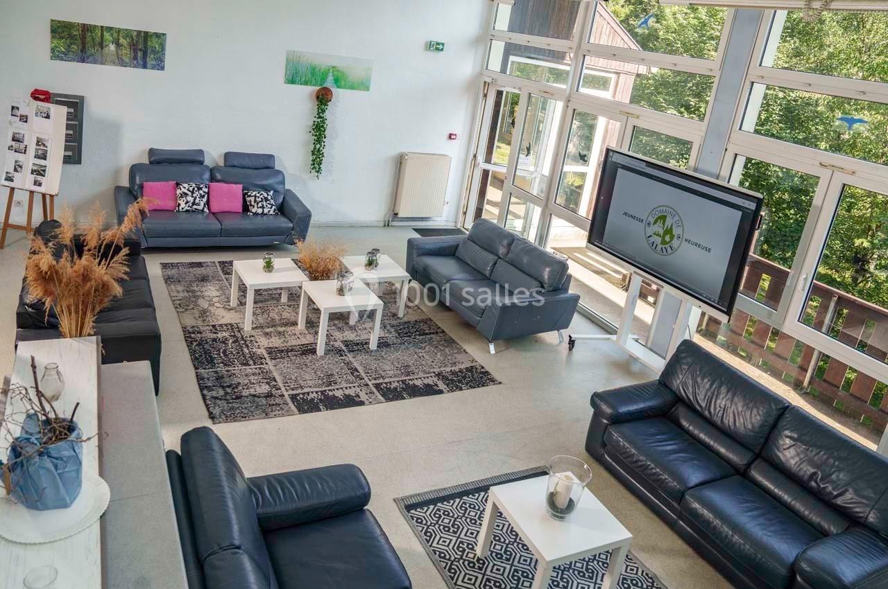 Salon lumineux avec canapés en cuir, tables basses, plantes décoratives et grande baie vitrée donnant sur un espace arboré.