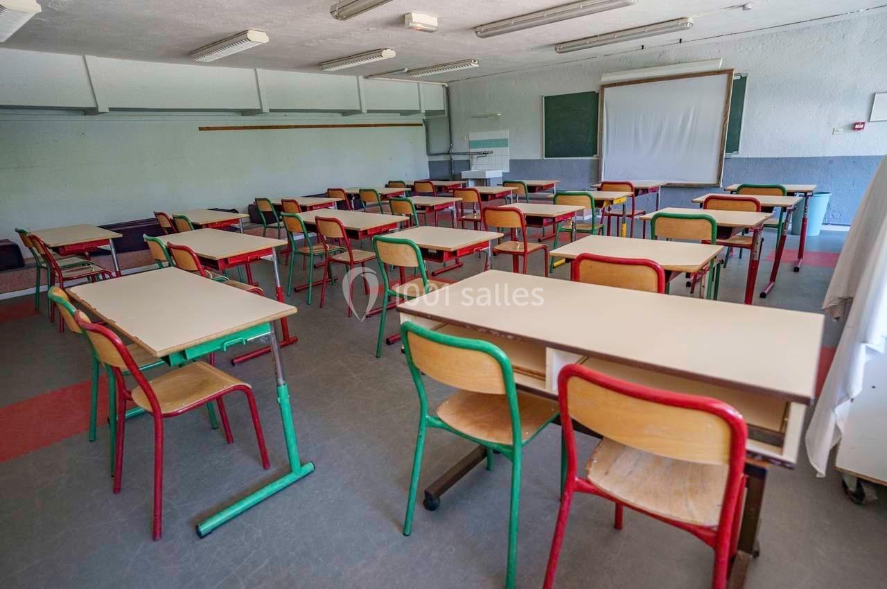 Salle de classe vide avec des bureaux et des chaises colorées alignés, tableau blanc et tableau noir au fond.