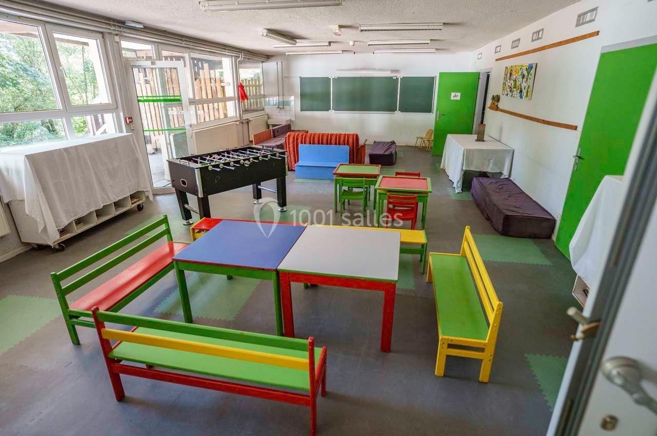 Salle de loisirs avec tables colorées, bancs, baby-foot et tableaux noirs, éclairée par de grandes fenêtres.