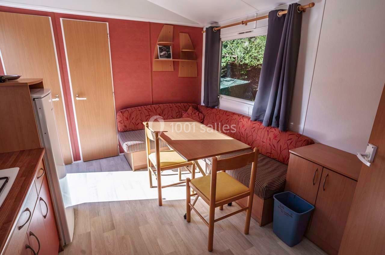 Intérieur d'un mobil-home avec coin salon, table en bois, chaises, fenêtre avec rideaux et rangements muraux.