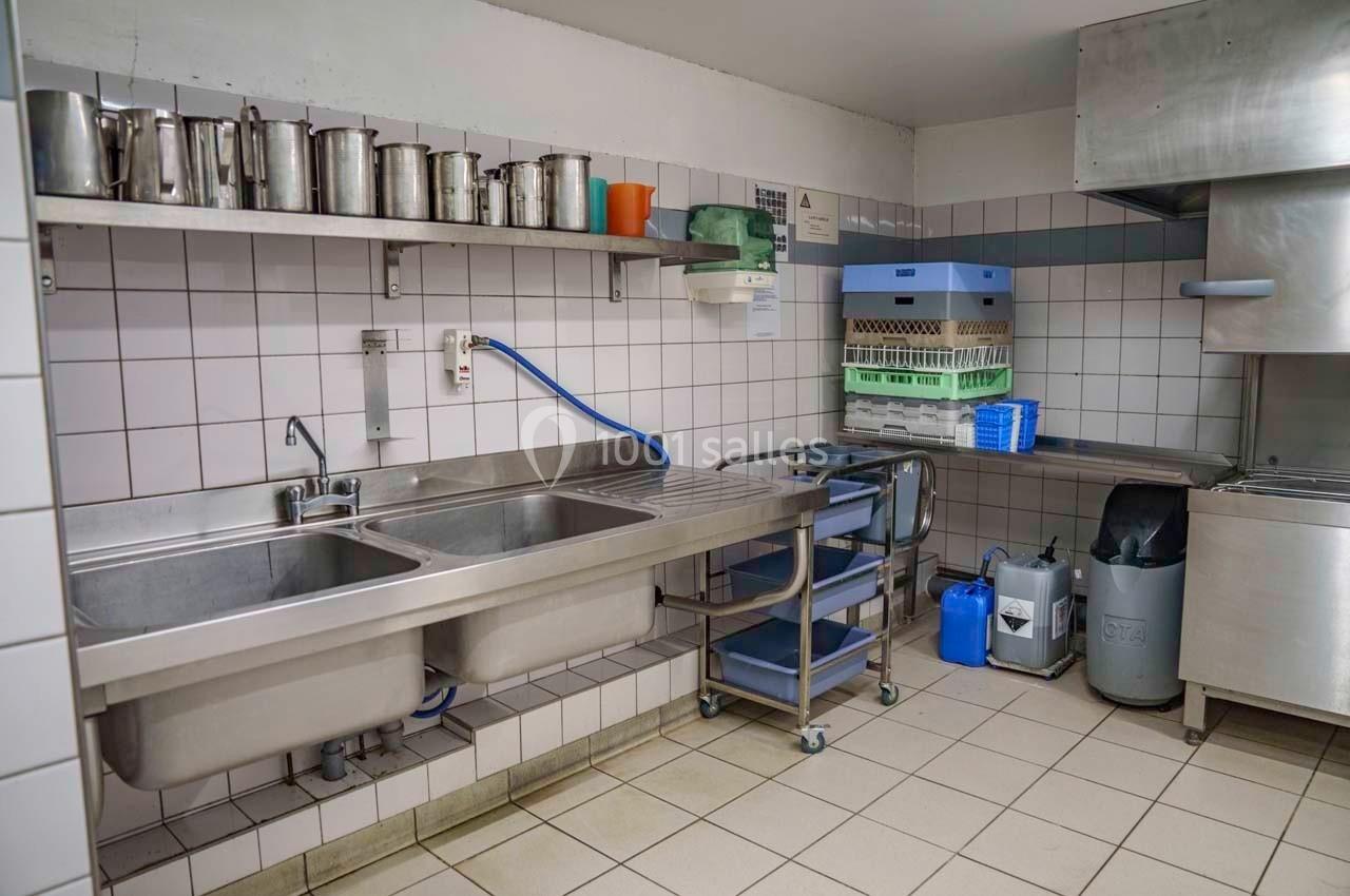 Cuisine professionnelle avec évier double, étagères métalliques, lave-vaisselle et carrelage mural blanc.