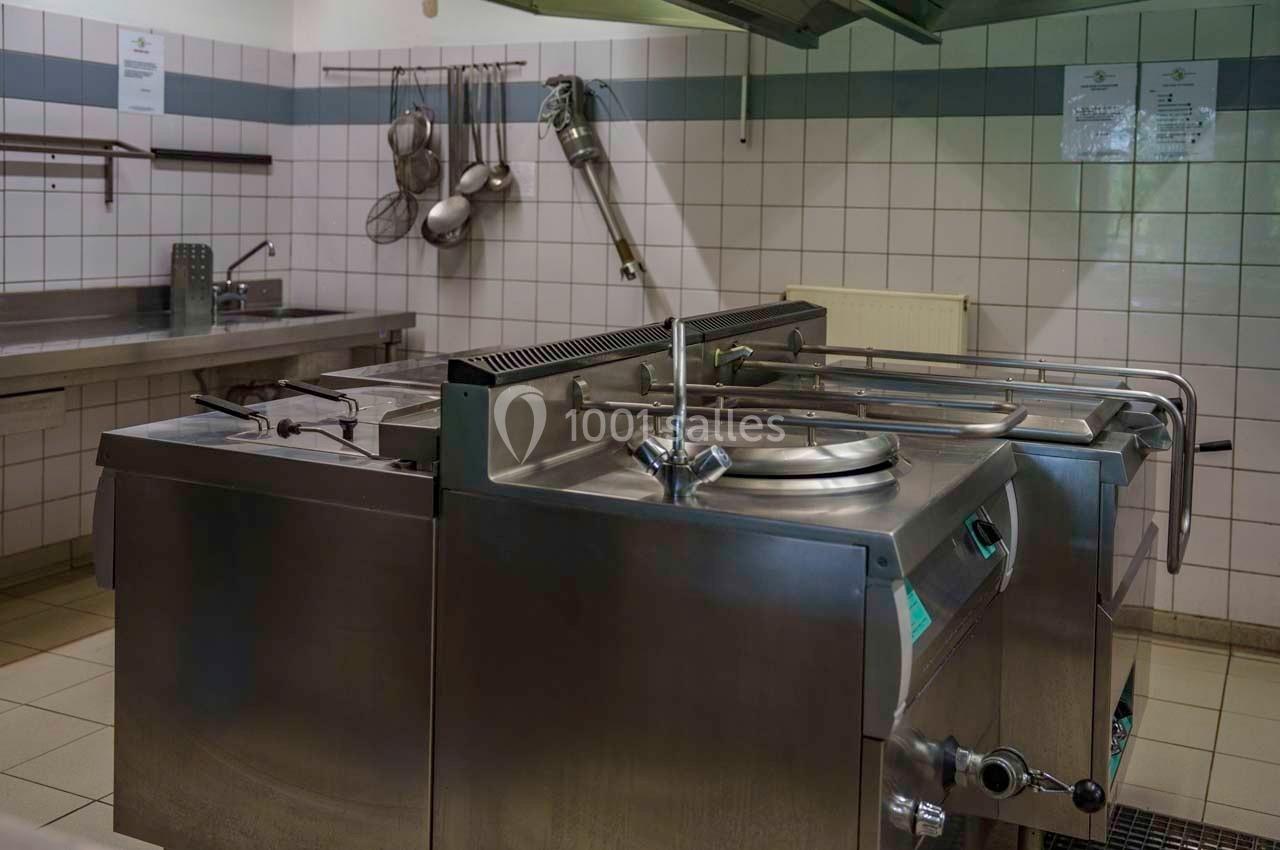 Cuisine professionnelle équipée de plaques de cuisson, marmites en inox et ustensiles suspendus sur un mur carrelé.