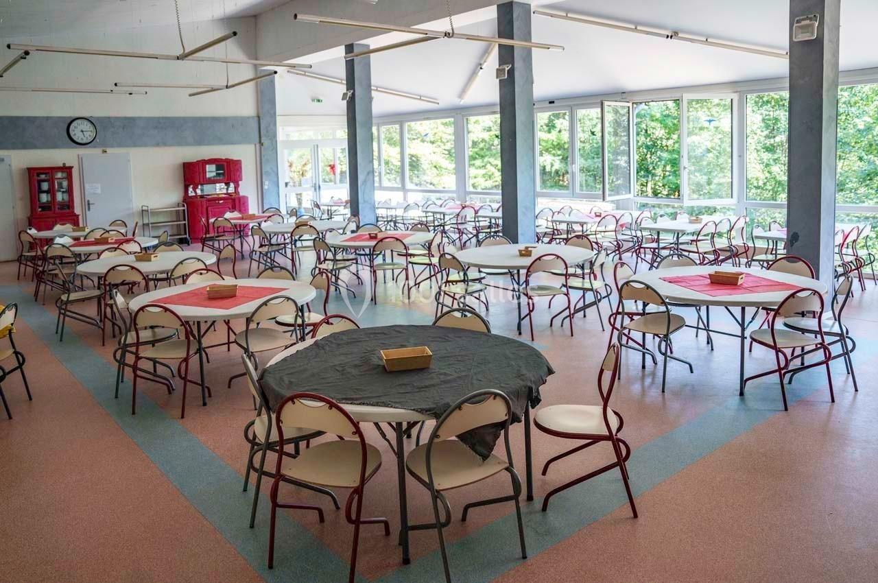 Salle lumineuse avec des tables rondes et chaises disposées pour un repas, grandes fenêtres donnant sur un espace vert.