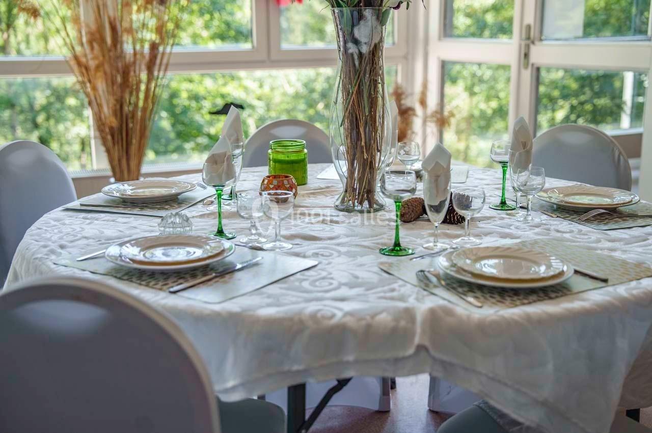 Table ronde dressée avec une nappe blanche, vaisselle, verres et décorations, devant des fenêtres donnant sur un jardin.