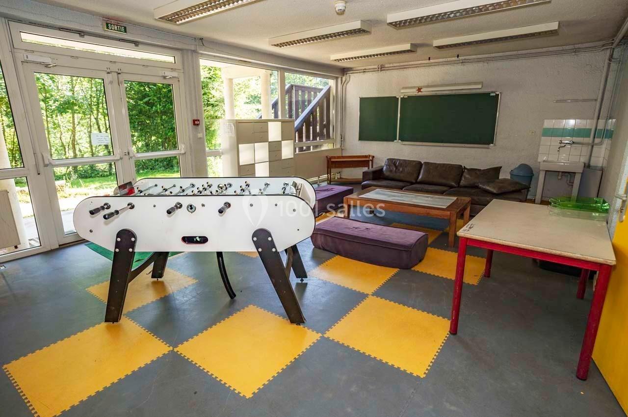 Salle de détente avec un baby-foot, des canapés, des tables et une vue sur l'extérieur verdoyant.