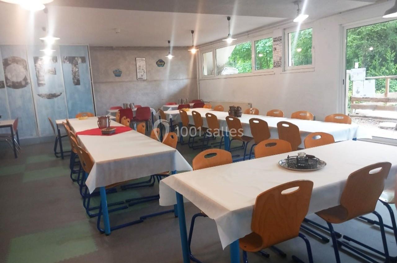 Salle lumineuse avec des tables alignées, nappées de blanc, entourées de chaises orange, et vue sur l'extérieur verdoyant.