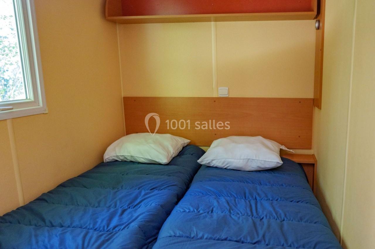 Deux lits simples avec couettes bleues dans une chambre aux murs beige, éclairée par une fenêtre à gauche.