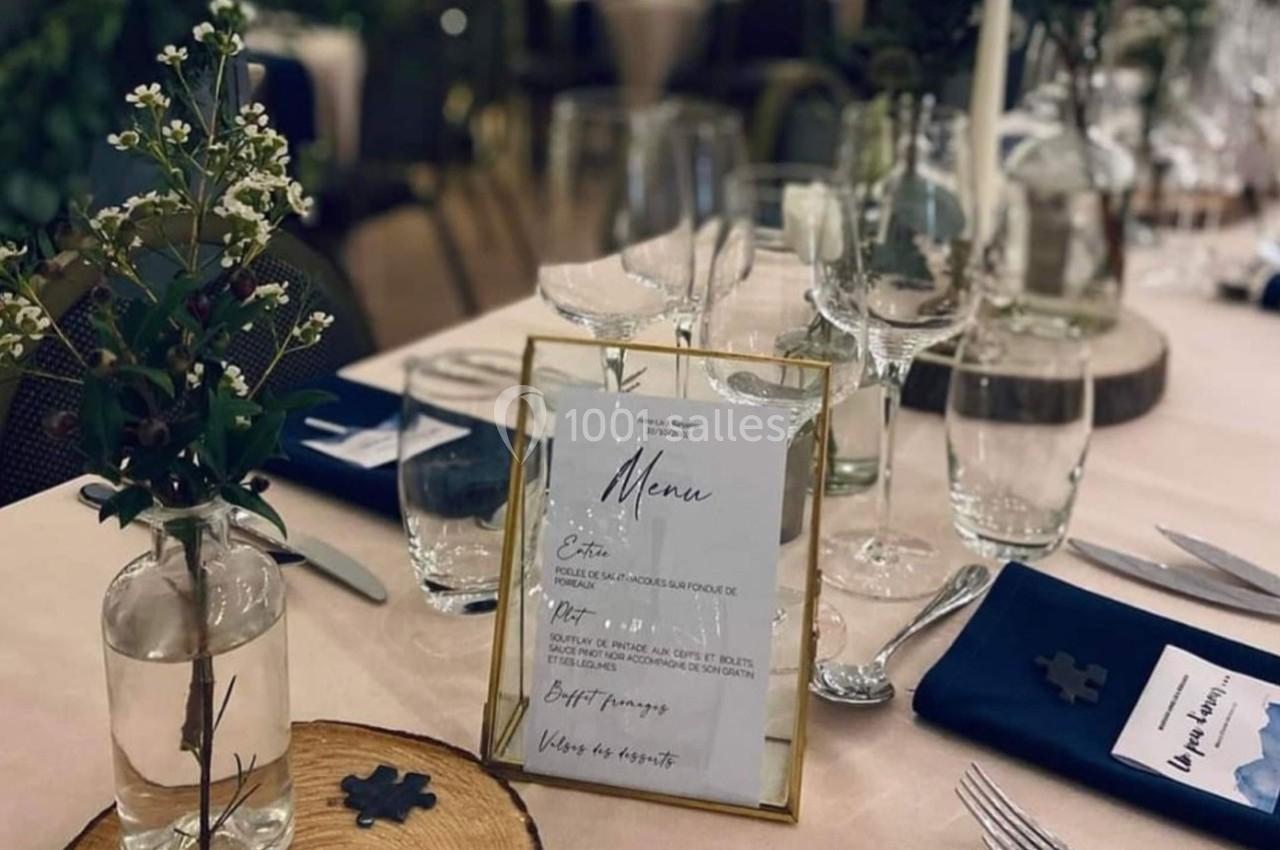 Table dressée avec menu encadré, verres, couverts, serviettes bleues et décoration florale sur nappe claire.