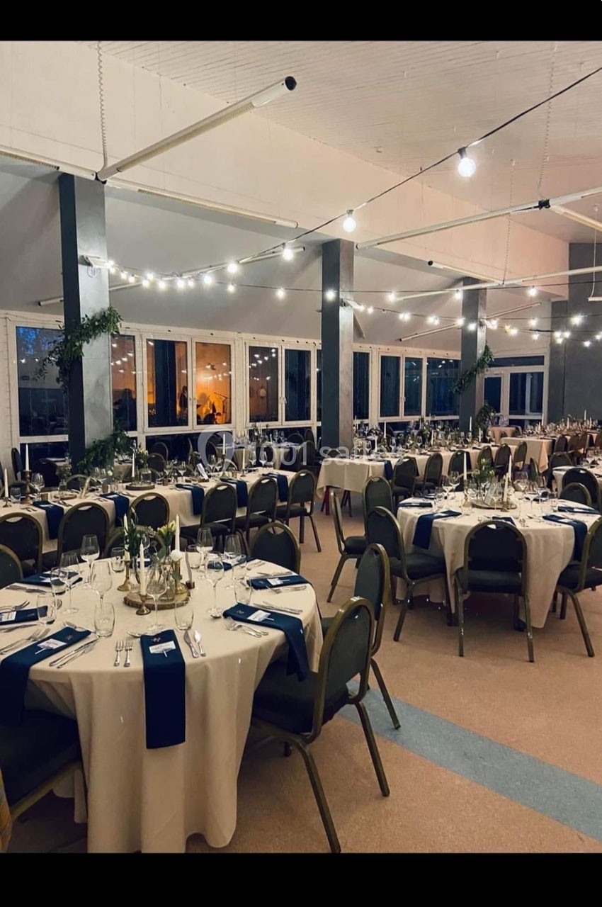 Salle de réception décorée avec des tables rondes dressées, nappes blanches et serviettes bleues, éclairage suspendu.