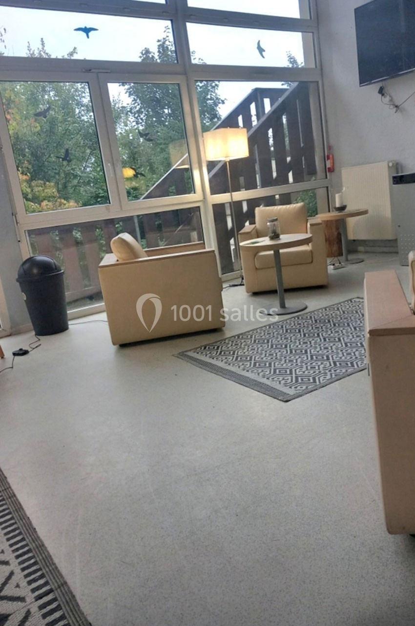 Salle lumineuse avec fauteuils beiges, tables basses, tapis, grande baie vitrée et vue sur des arbres.