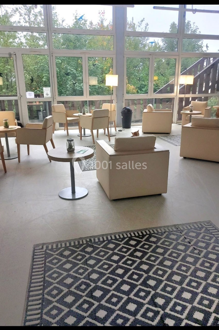 Espace lumineux avec fauteuils beiges, tables basses et grandes baies vitrées donnant sur un environnement arboré.