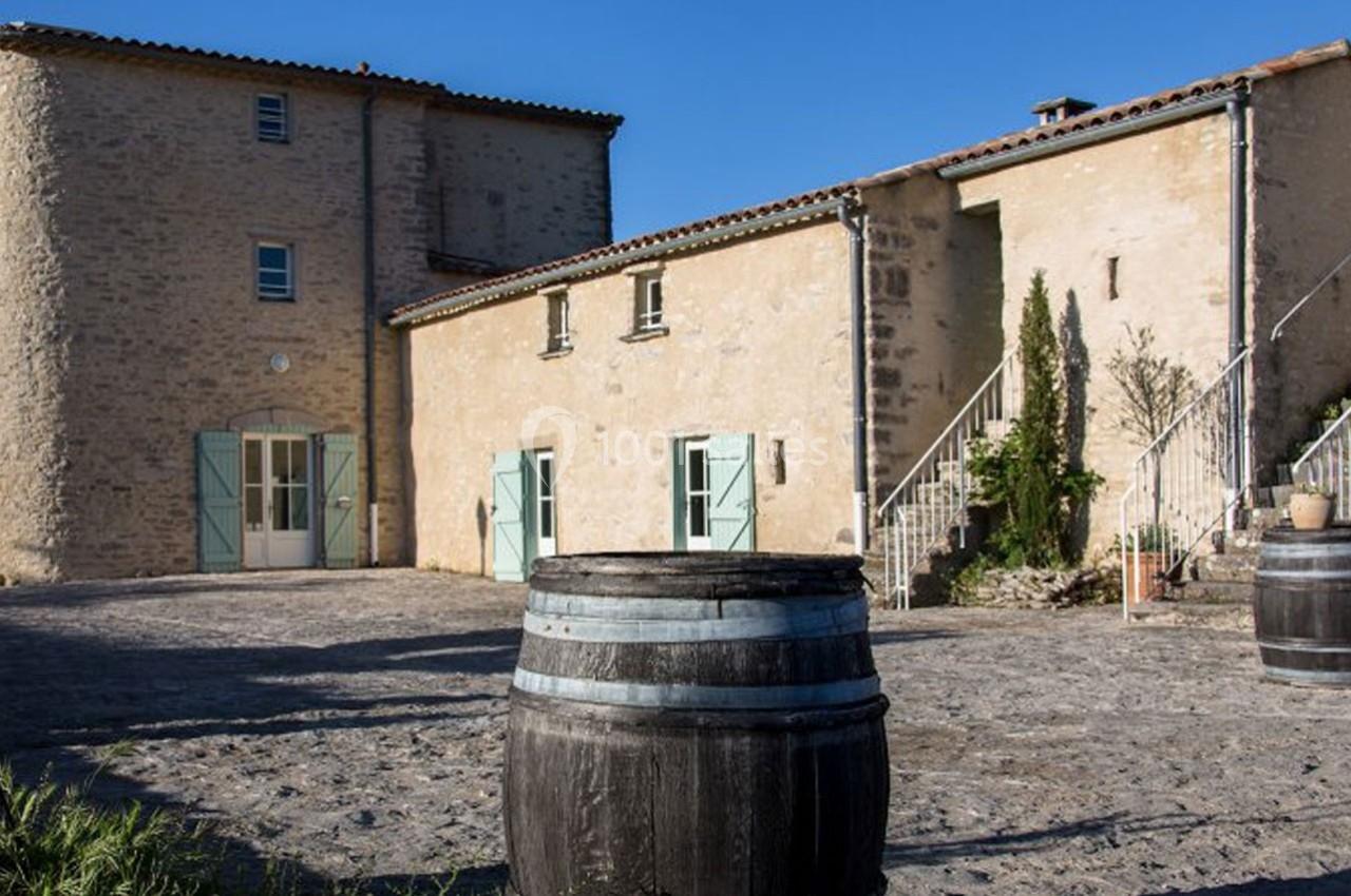 Cour pavée avec des bâtiments en pierre, volets pastel, barils en bois et escalier extérieur sous un ciel bleu.