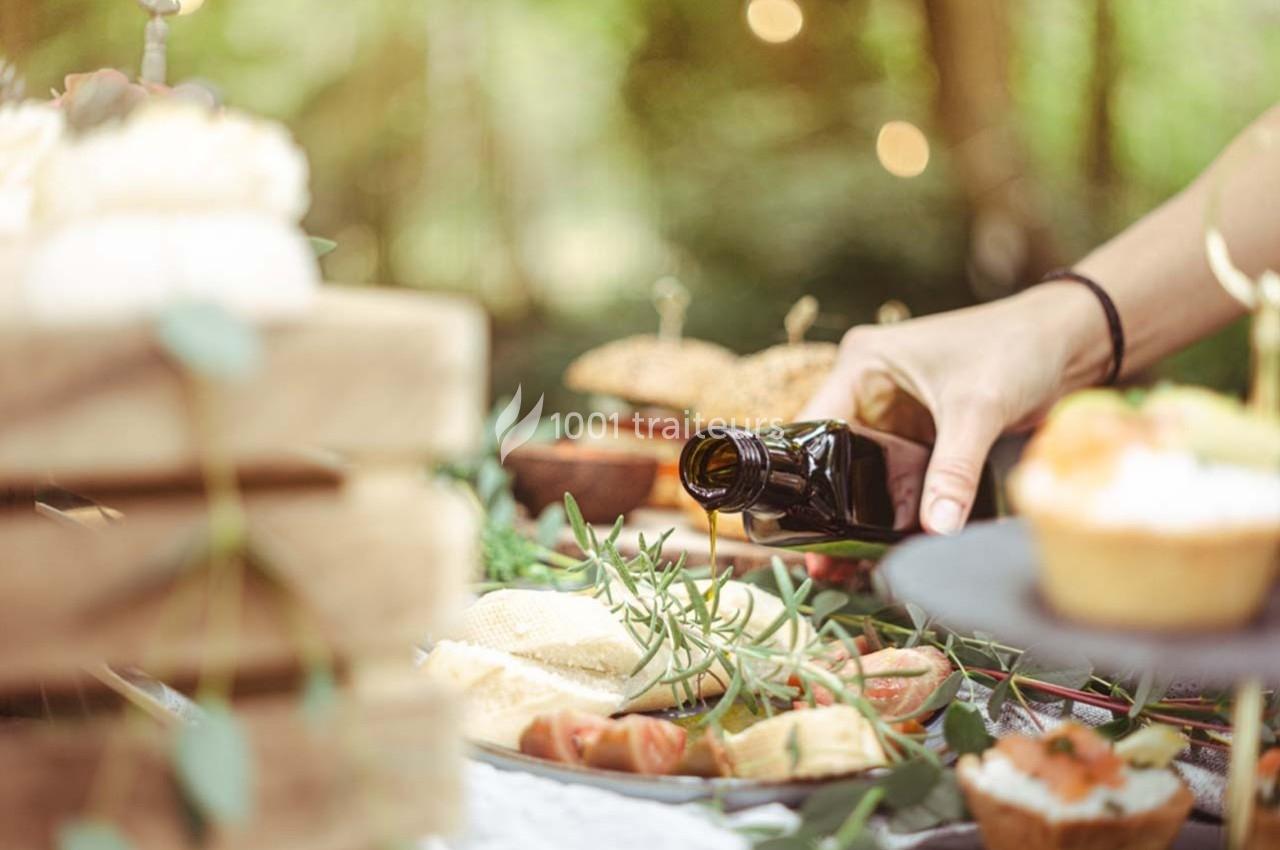 Une main verse de l'huile d'olive sur une table garnie de plats et de garnitures dans un cadre extérieur verdoyant.