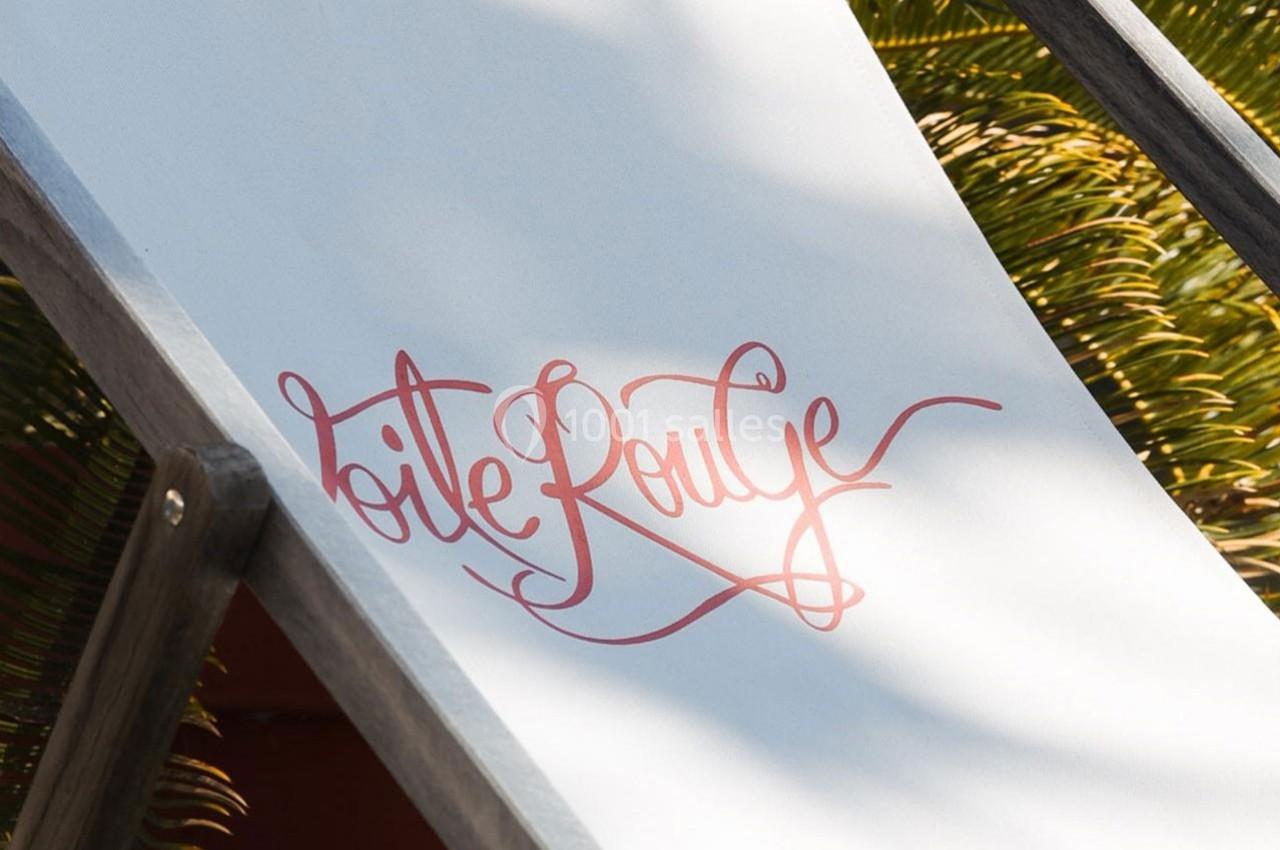 Texte ’Toile Rouge’ écrit en rouge sur une toile blanche d'un transat, avec des feuilles de palmier en arrière-plan.