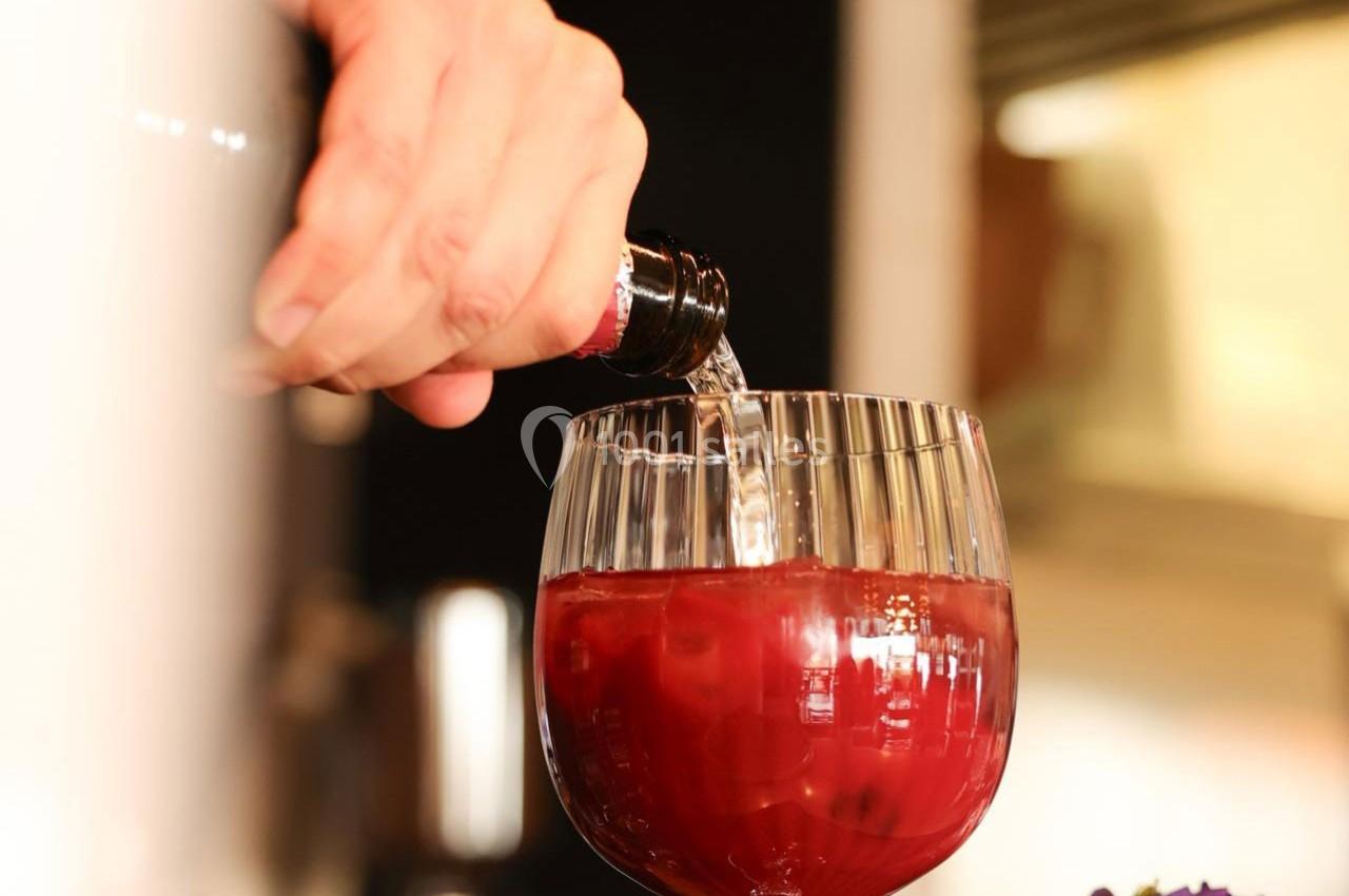Main versant une boisson transparente dans un verre rempli d'un liquide rouge, posé sur une table.