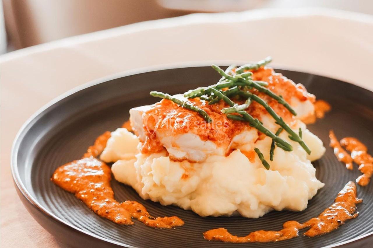 Plat de poisson sur purée de pommes de terre, nappé de sauce rouge et garni de salicornes, présenté dans une assiette noire.