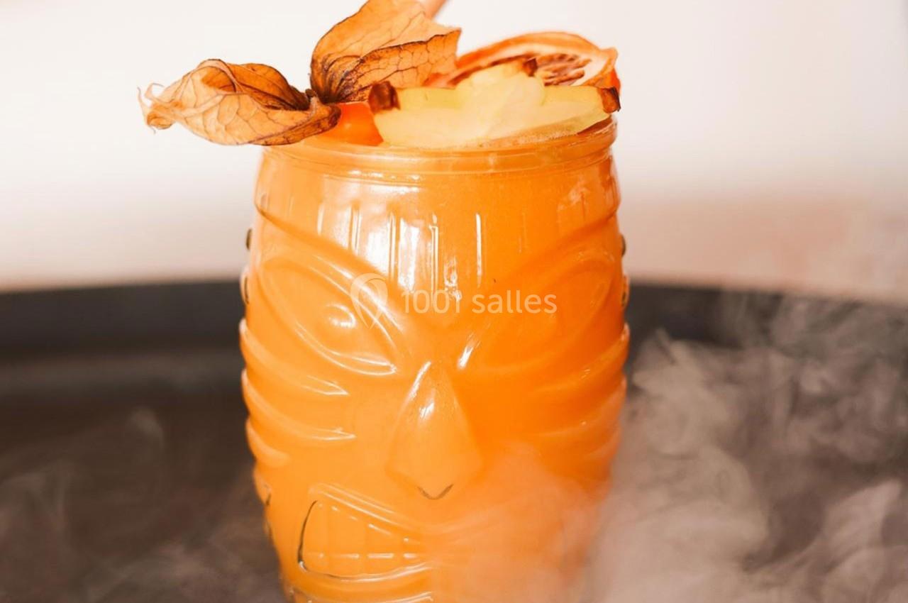 Cocktail orange servi dans un verre tiki décoré de physalis, ananas et fumée en arrière-plan.