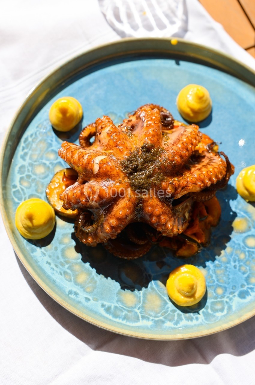 Assiette bleue contenant un poulpe grillé entouré de touches de sauce jaune, présentée sur une nappe blanche.