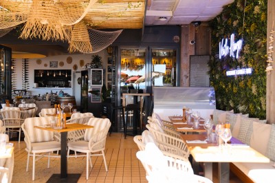 Salle de restaurant lumineuse avec tables dressées, chaises rembourrées, mur décoré de bouteilles de vin et lustre suspendu.