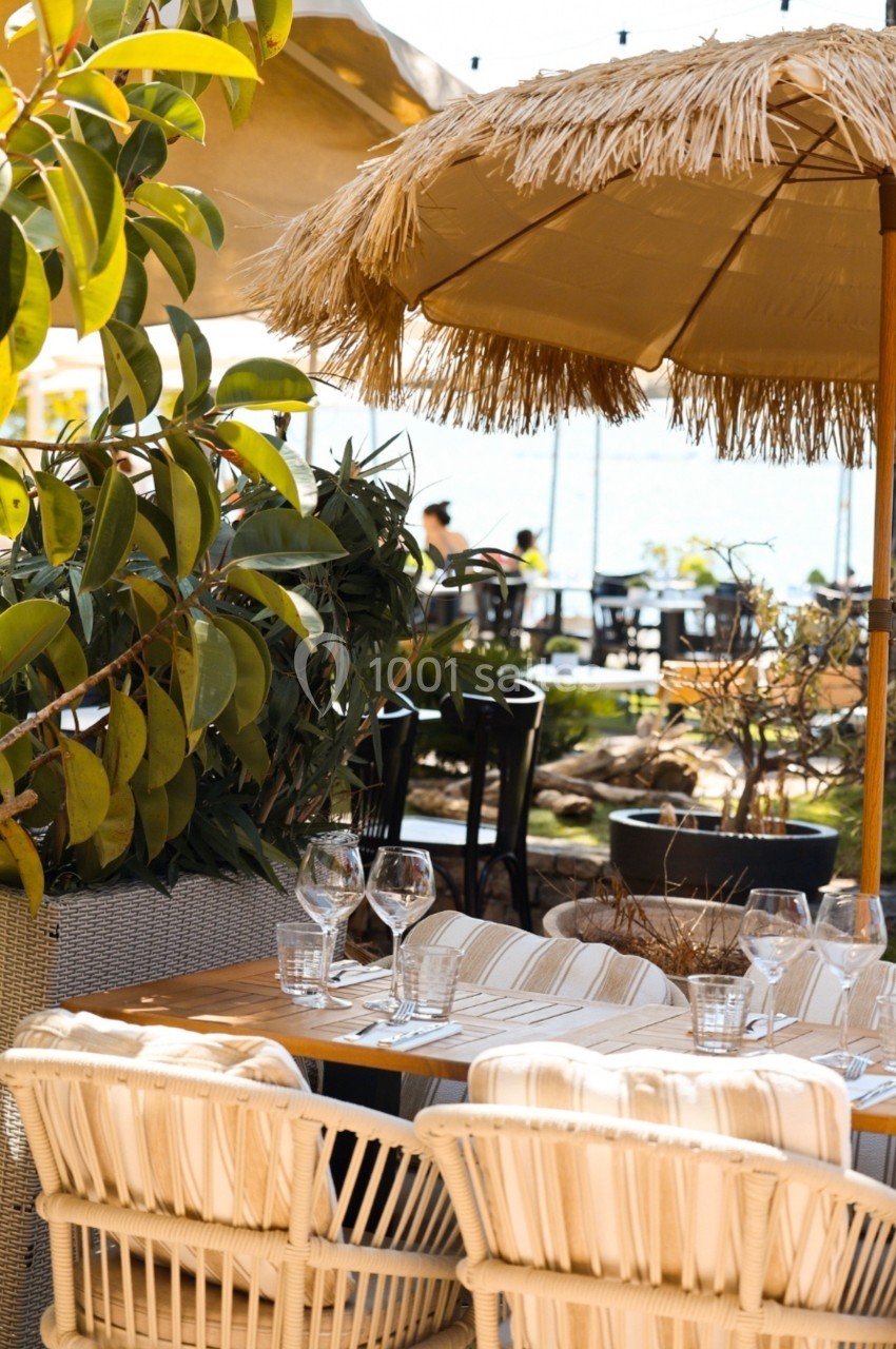 Table en rotin dressée avec des verres et des assiettes, sous un parasol en paille dans un jardin ensoleillé.