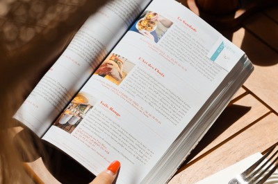 Salle de restaurant lumineuse avec tables dressées, chaises rembourrées, mur décoré de bouteilles de vin et lustre suspendu.
