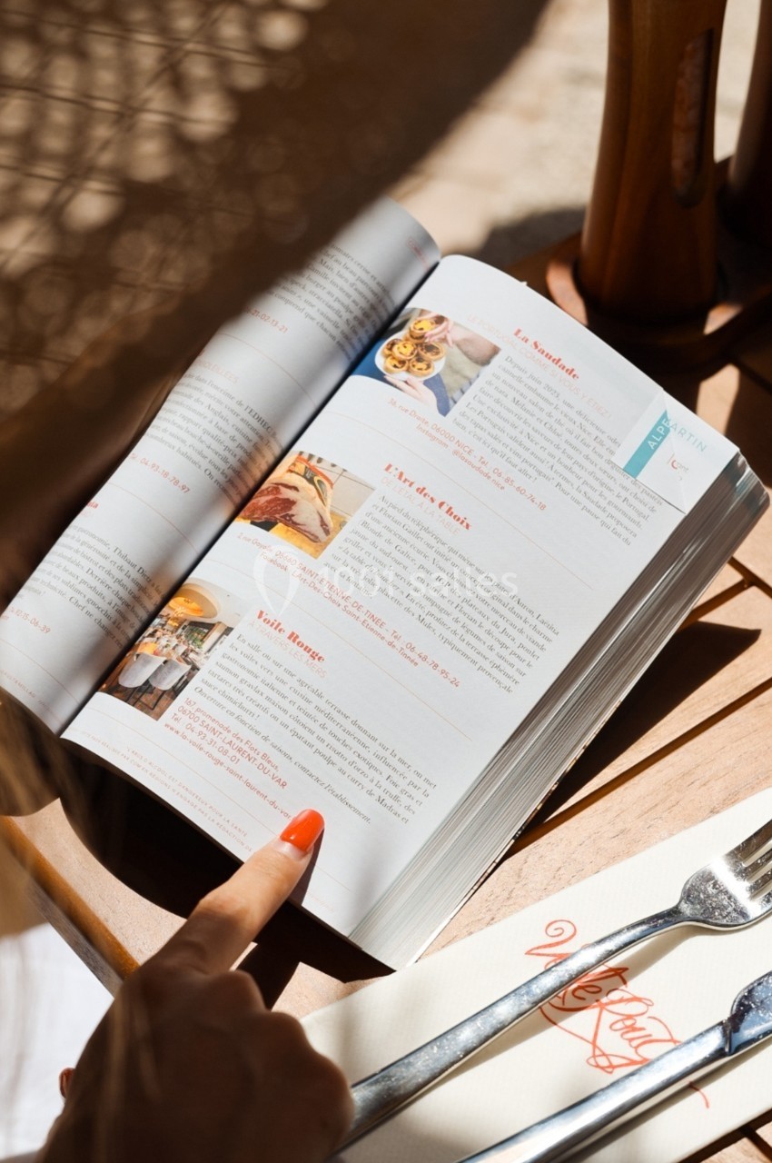 Une personne lit un magazine ouvert sur une table en bois, avec des couverts et une serviette à proximité.