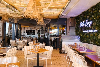 Salle de restaurant lumineuse avec tables dressées, chaises rembourrées, mur décoré de bouteilles de vin et lustre suspendu.