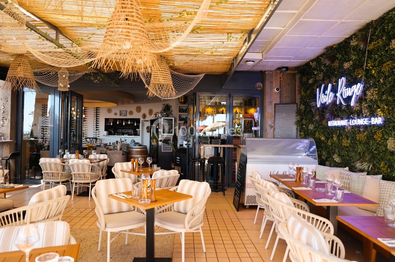 Terrasse d'un restaurant avec mobilier en bois et rotin, décoration végétale et enseigne lumineuse sur un mur.