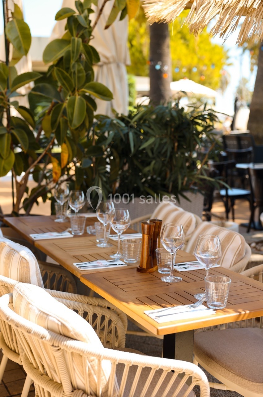 Table en bois dressée avec verres et couverts, entourée de chaises en rotin, dans un cadre extérieur végétalisé.