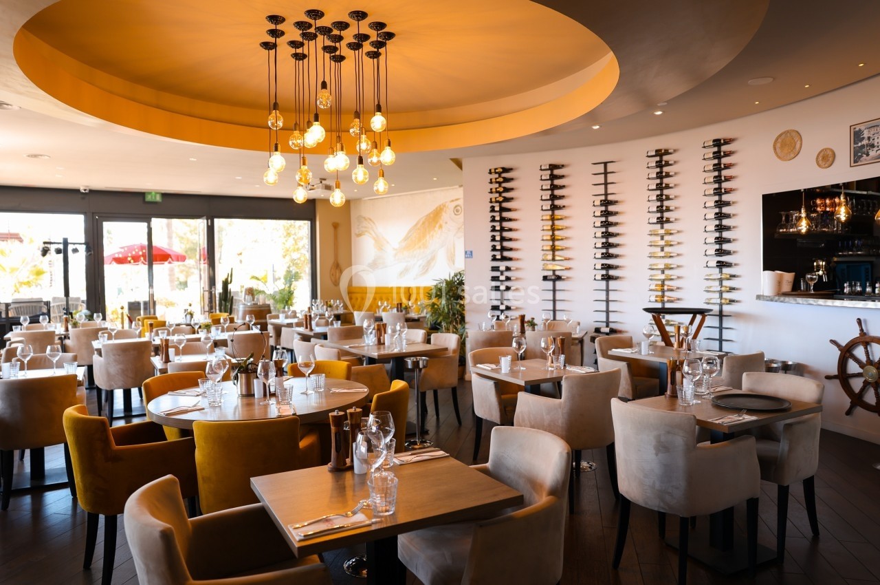 Salle de restaurant lumineuse avec tables dressées, chaises rembourrées, mur décoré de bouteilles de vin et lustre suspendu.