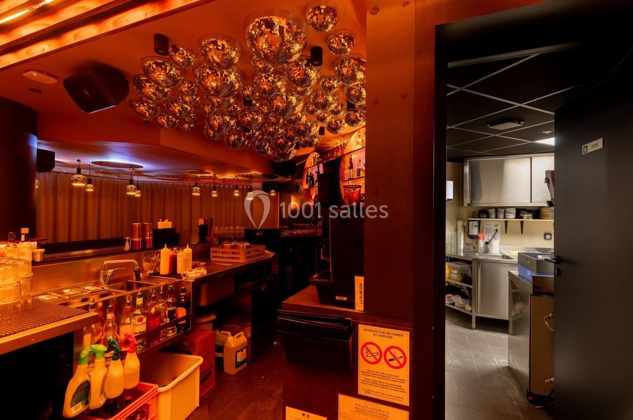 Intérieur d'un bar avec comptoir, bouteilles, éclairage tamisé et vue sur une cuisine professionnelle à droite.