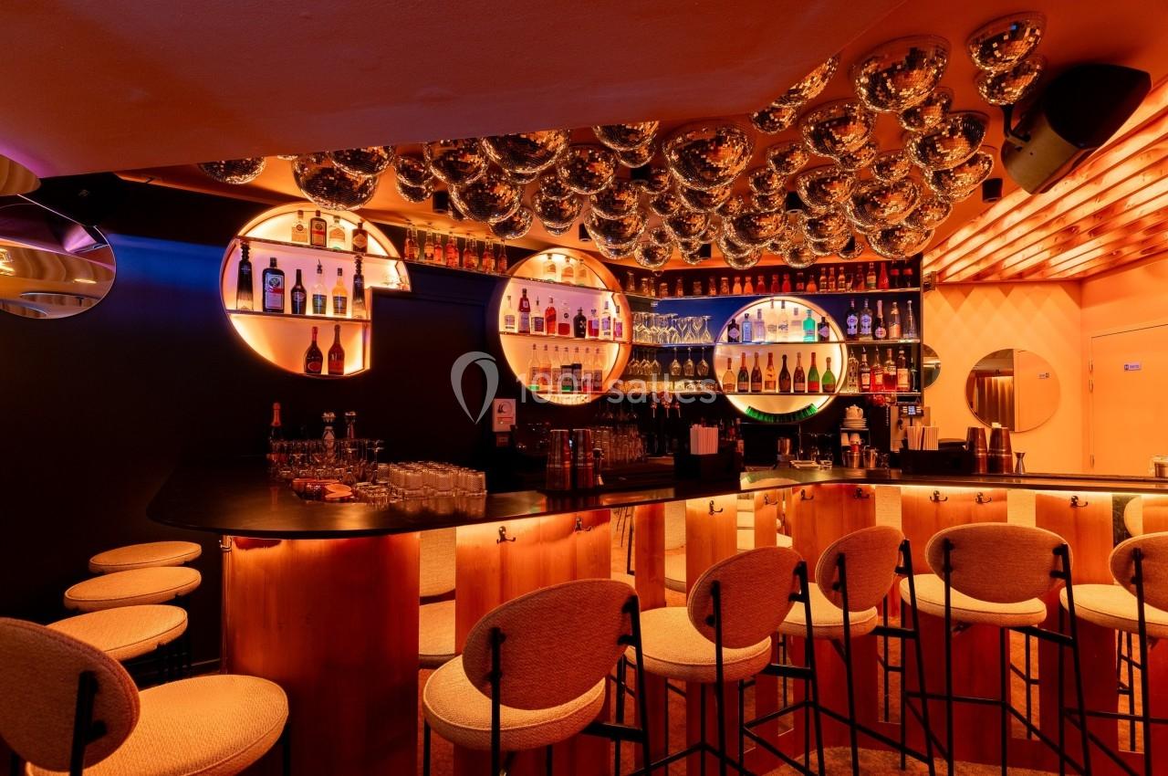 Bar moderne avec comptoir en bois, tabourets rembourrés, étagères éclairées contenant des bouteilles et décorations…