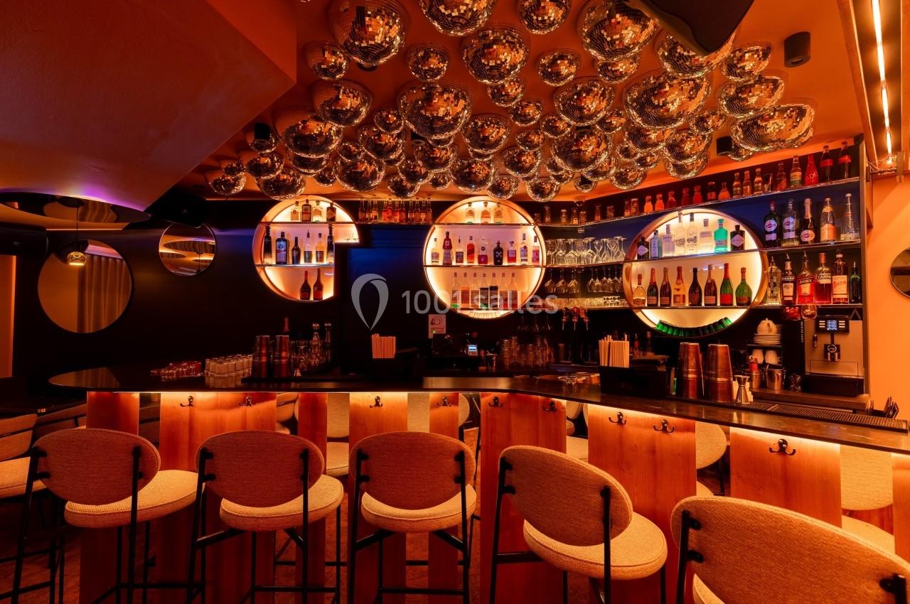 Bar avec comptoir en bois, tabourets rembourrés, étagères de bouteilles éclairées et plafond décoré de sphères métalliques.