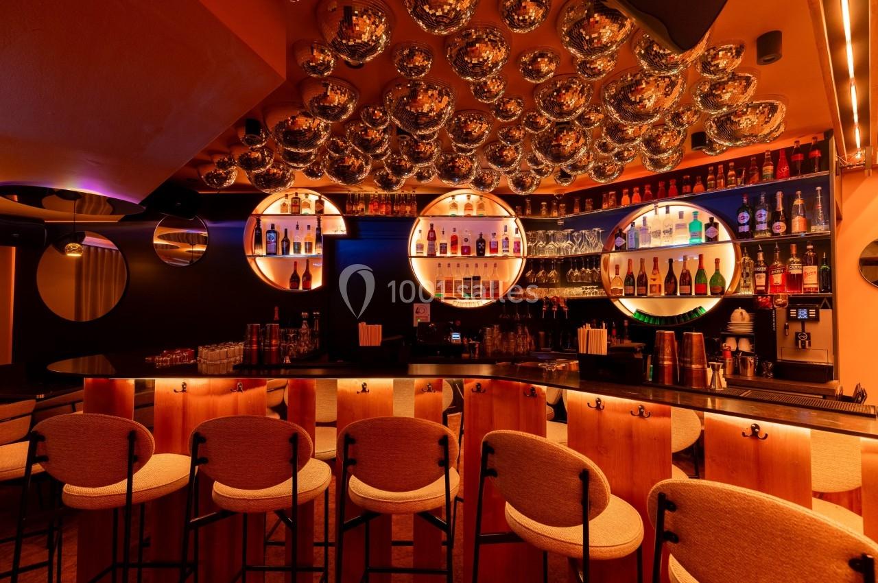 Bar moderne avec comptoir en bois, tabourets rembourrés, étagères illuminées de bouteilles et plafond décoré de sphères…