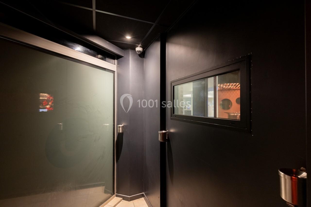 Vue d'un couloir sombre avec une porte vitrée givrée à gauche et une fenêtre rectangulaire sur un mur noir à droite.