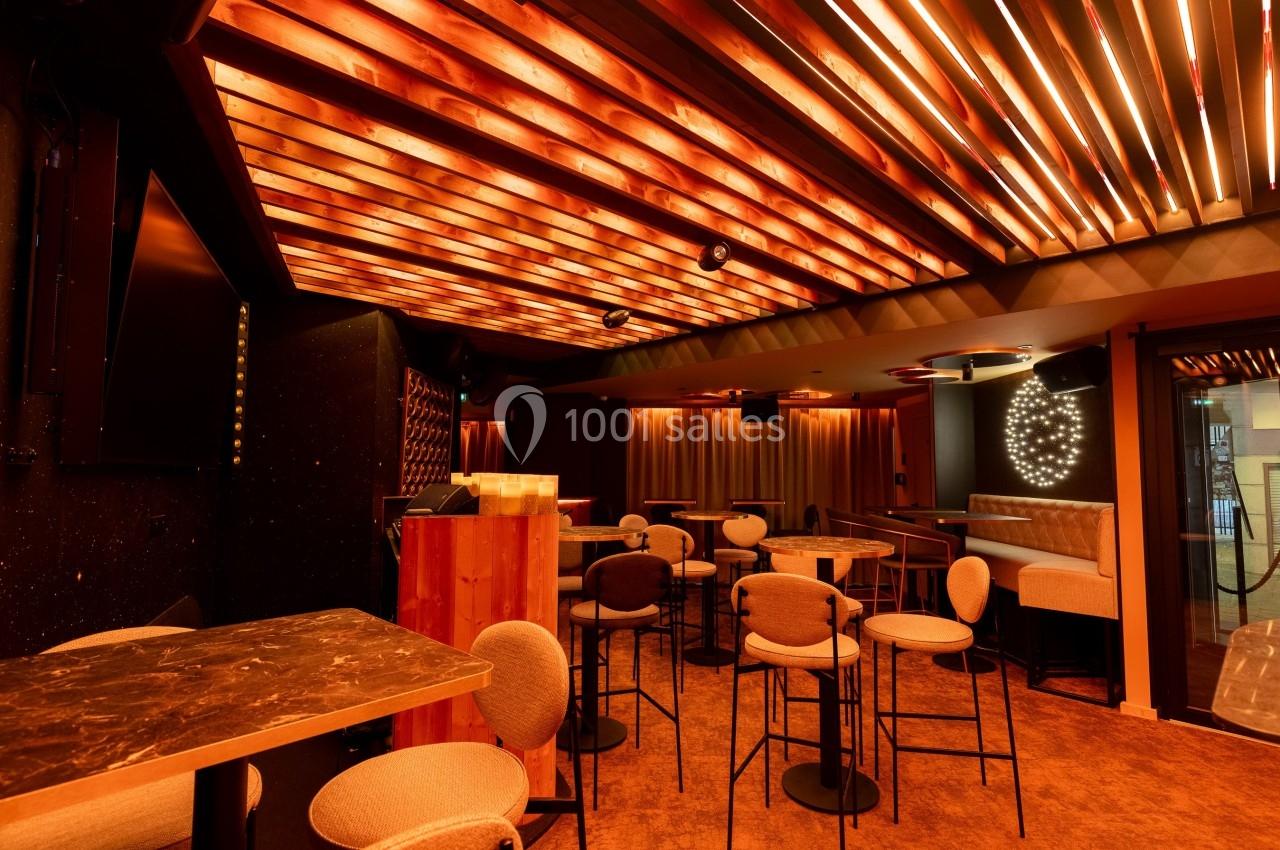 Salle moderne avec éclairage chaleureux, mobilier en bois et métal, tables hautes et banquettes dans un décor intimiste.