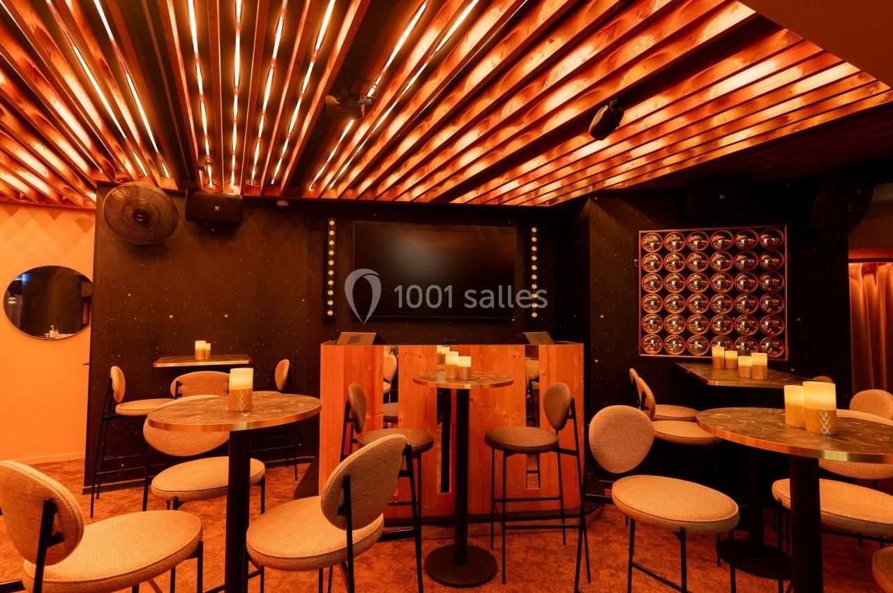 Salle avec tables hautes, chaises rembourrées, éclairage chaleureux au plafond et écran noir sur un mur sombre.