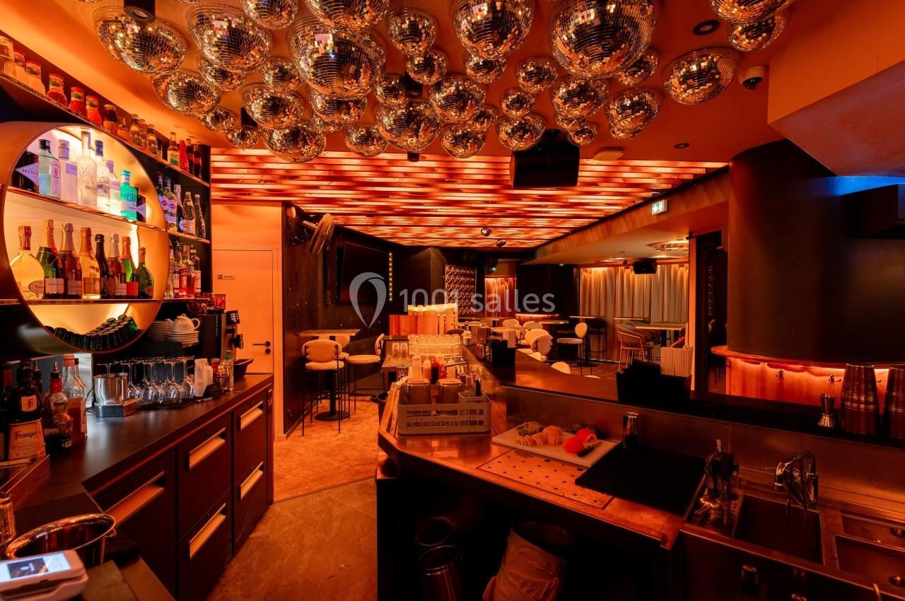 Intérieur d'un bar avec comptoir, étagères de bouteilles, tables, chaises et plafond décoré de boules à facettes.