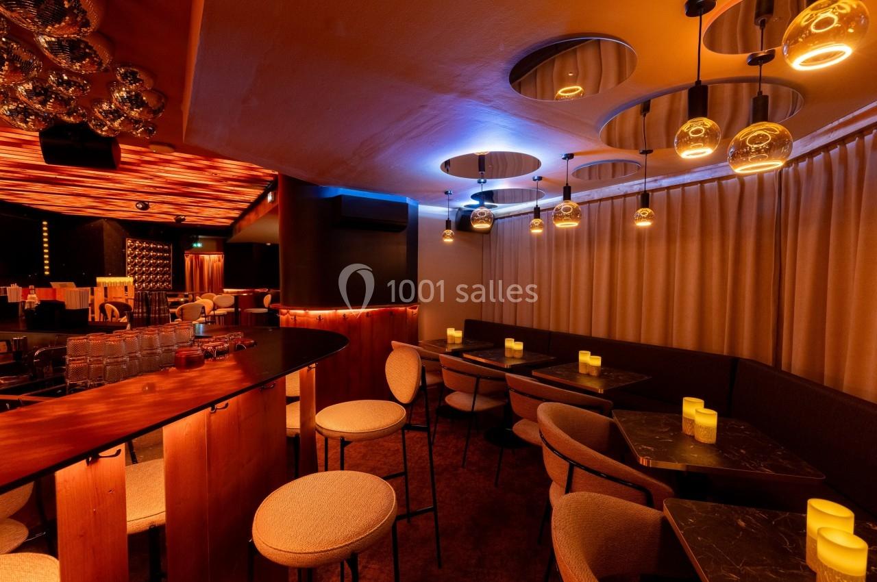 Salle de bar avec tables, chaises rembourrées, éclairage chaleureux et plafonds décorés de miroirs et luminaires.