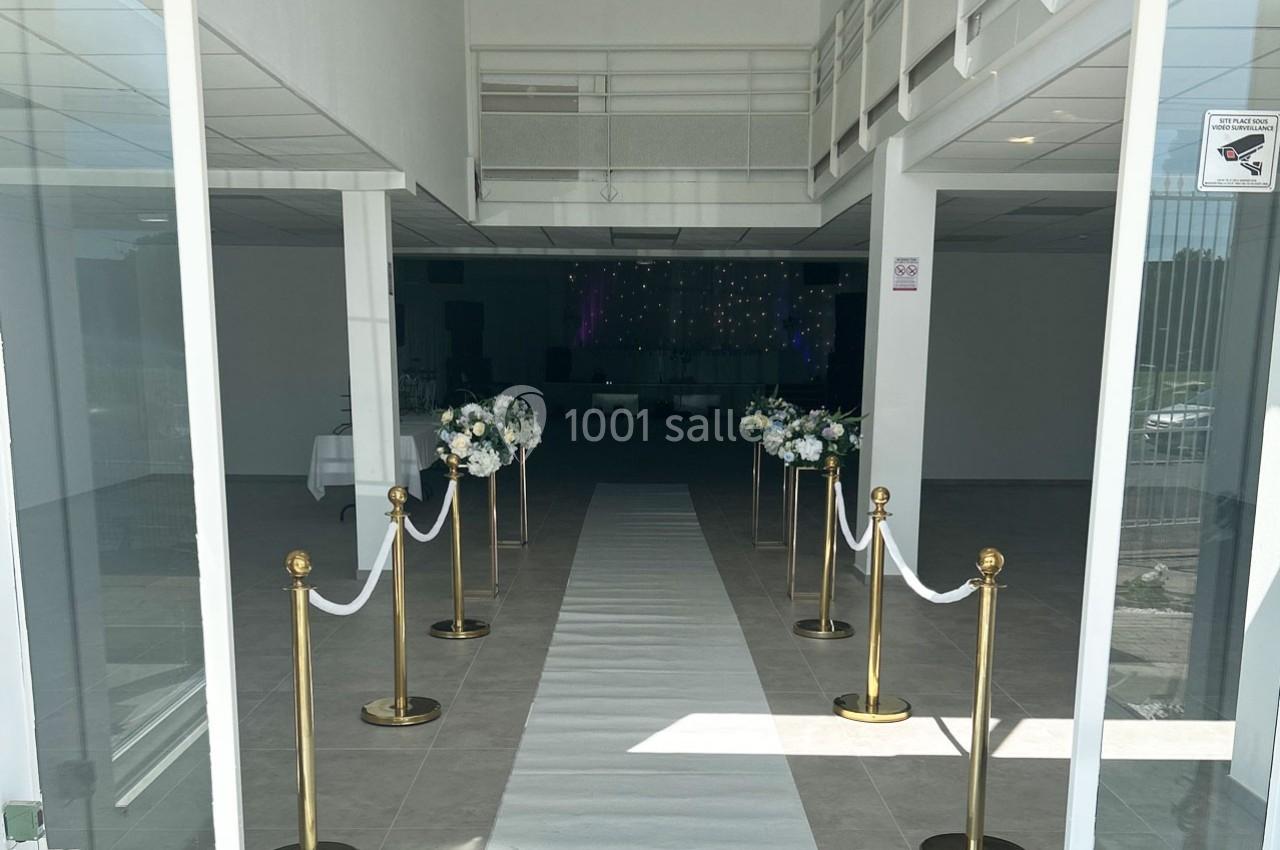 Salle de réception lumineuse avec allée centrale bordée de poteaux dorés, cordons blancs et arrangements floraux.