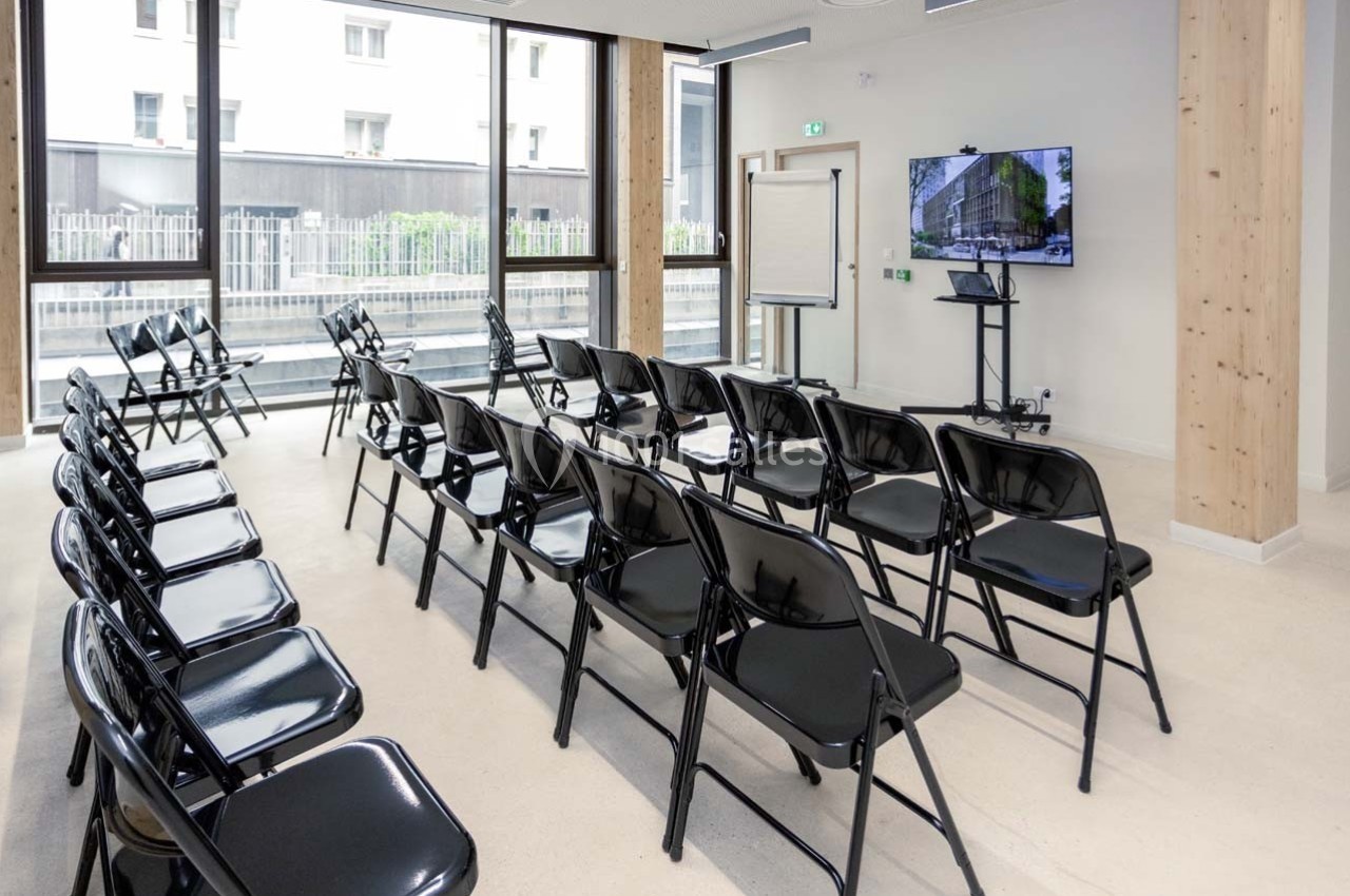 Salle de réunion lumineuse avec des chaises pliantes noires disposées en rangées face à un écran de présentation.