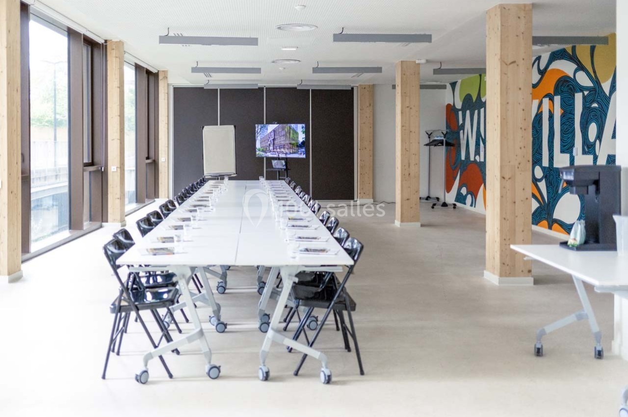 Salle de réunion lumineuse avec une longue table, des chaises noires, un écran, un tableau blanc et une fresque murale…