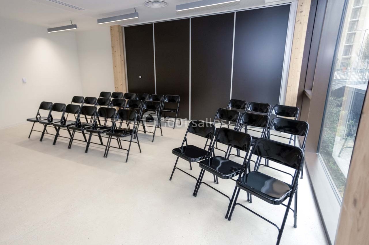 Salle lumineuse avec rangées de chaises pliantes noires alignées face à un mur sombre et une grande fenêtre.