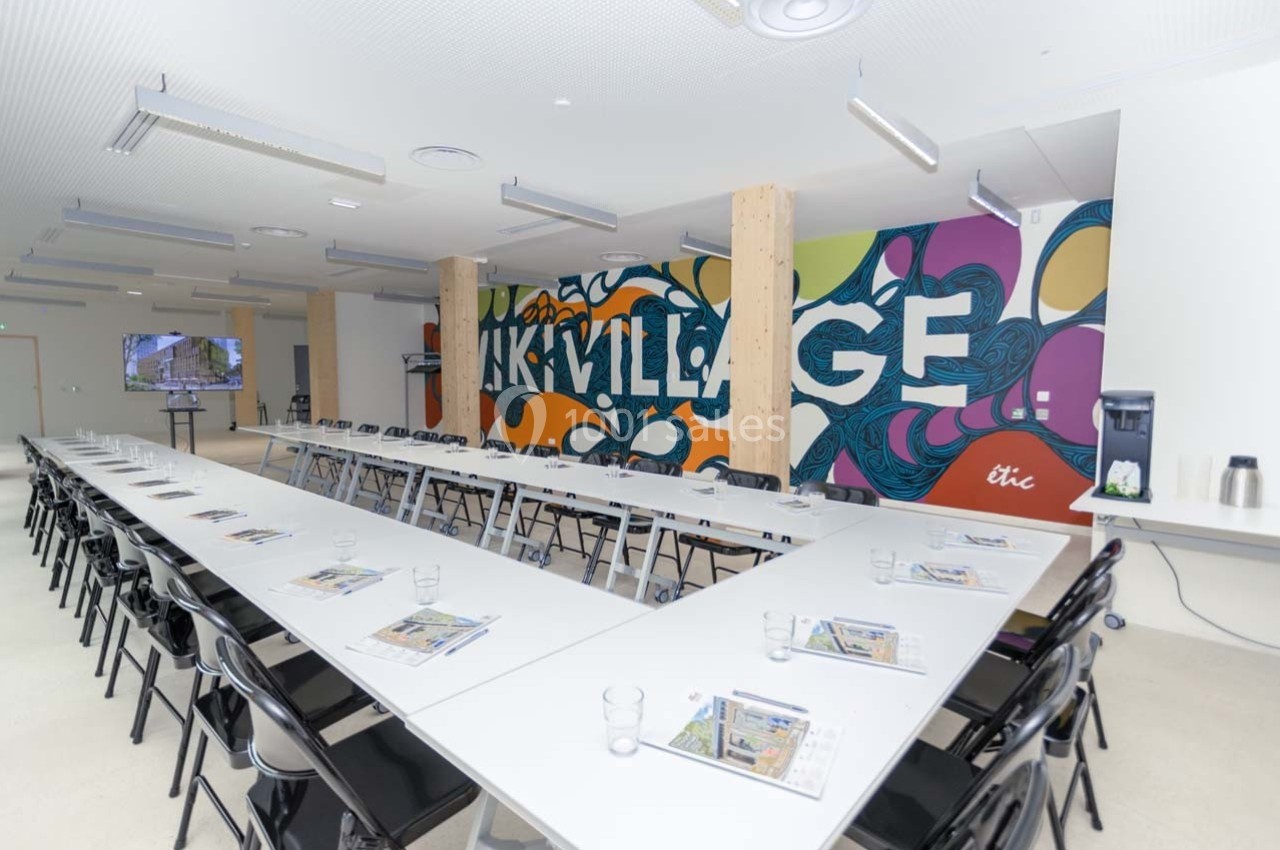 Salle de réunion lumineuse avec de longues tables en U, chaises noires, mur coloré et distributeur d'eau.
