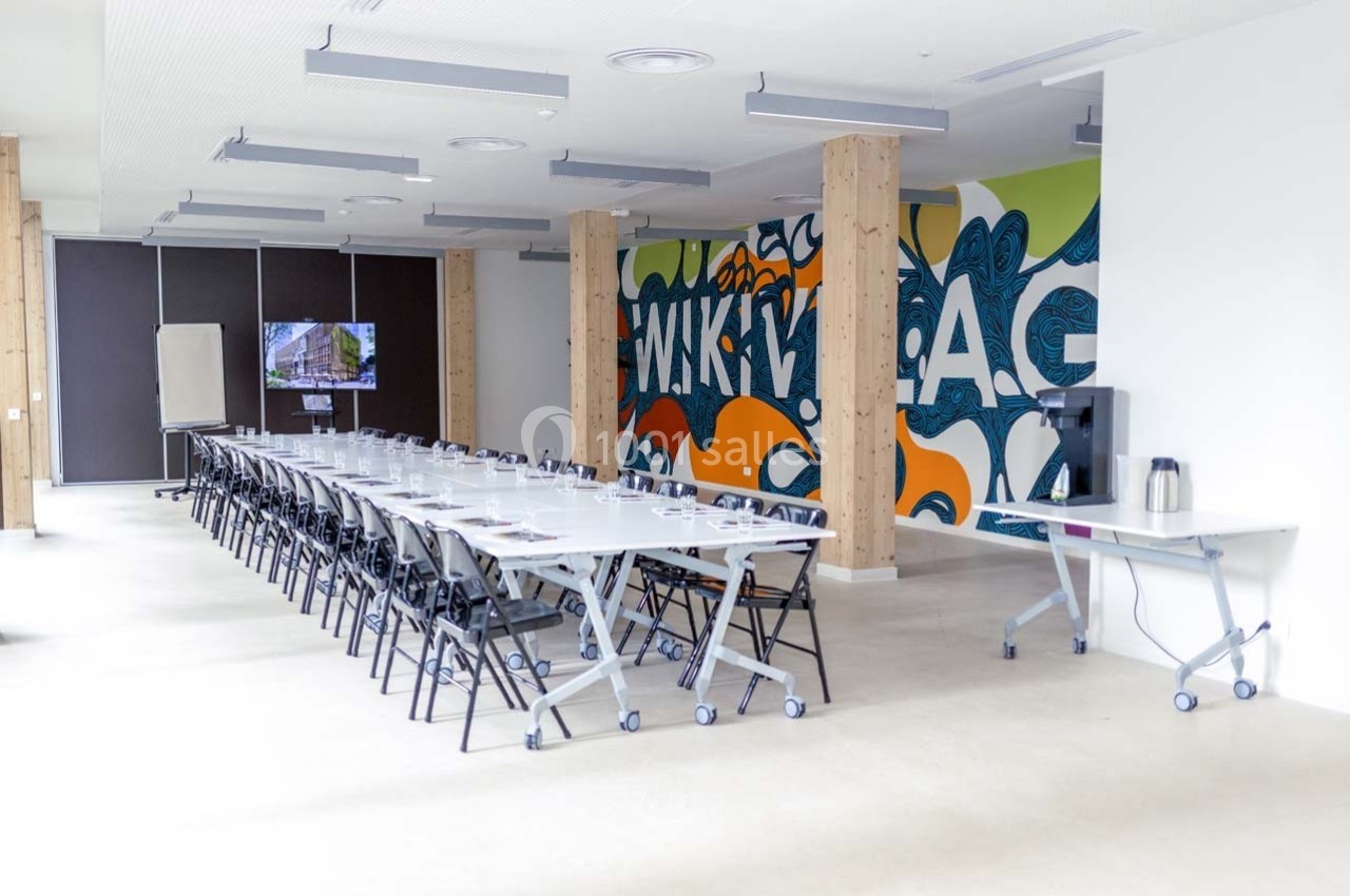 Salle de réunion lumineuse avec longues tables, chaises pliantes, mur coloré et écran de présentation.
