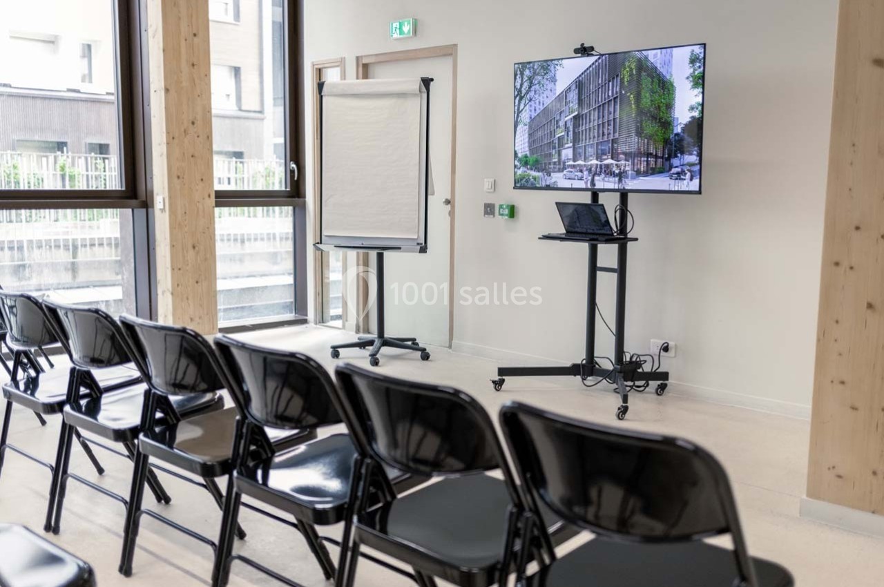 Salle de réunion avec chaises noires alignées, écran affichant un bâtiment et un tableau de conférence sur pied.