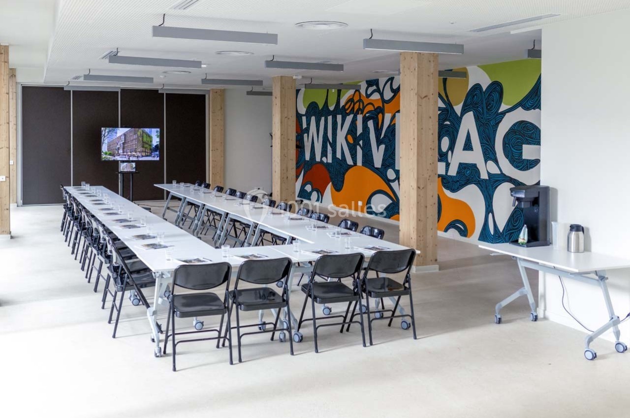 Salle de réunion lumineuse avec longues tables, chaises noires, mur décoré de graffitis et écran de présentation.