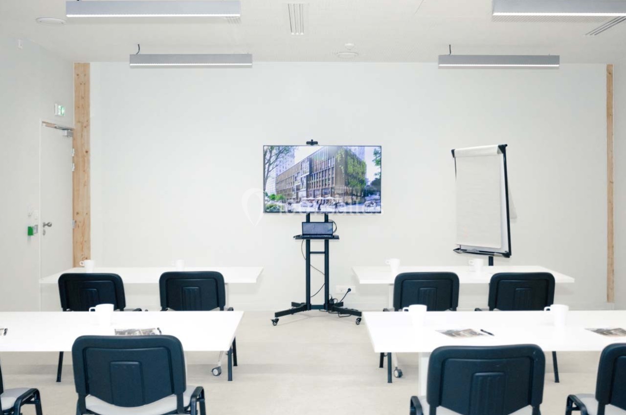 Salle de réunion avec des tables, des chaises, un écran affichant un bâtiment, un chevalet et des gobelets disposés.