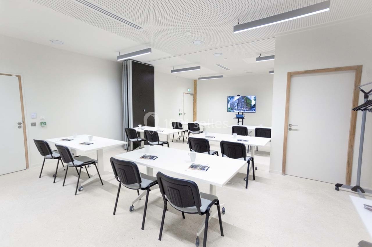 Salle de formation lumineuse avec des tables blanches, des chaises noires et un écran affichant une image.